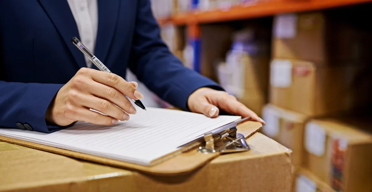 A person checking a clipboard against stacked export cartons in a warehouse